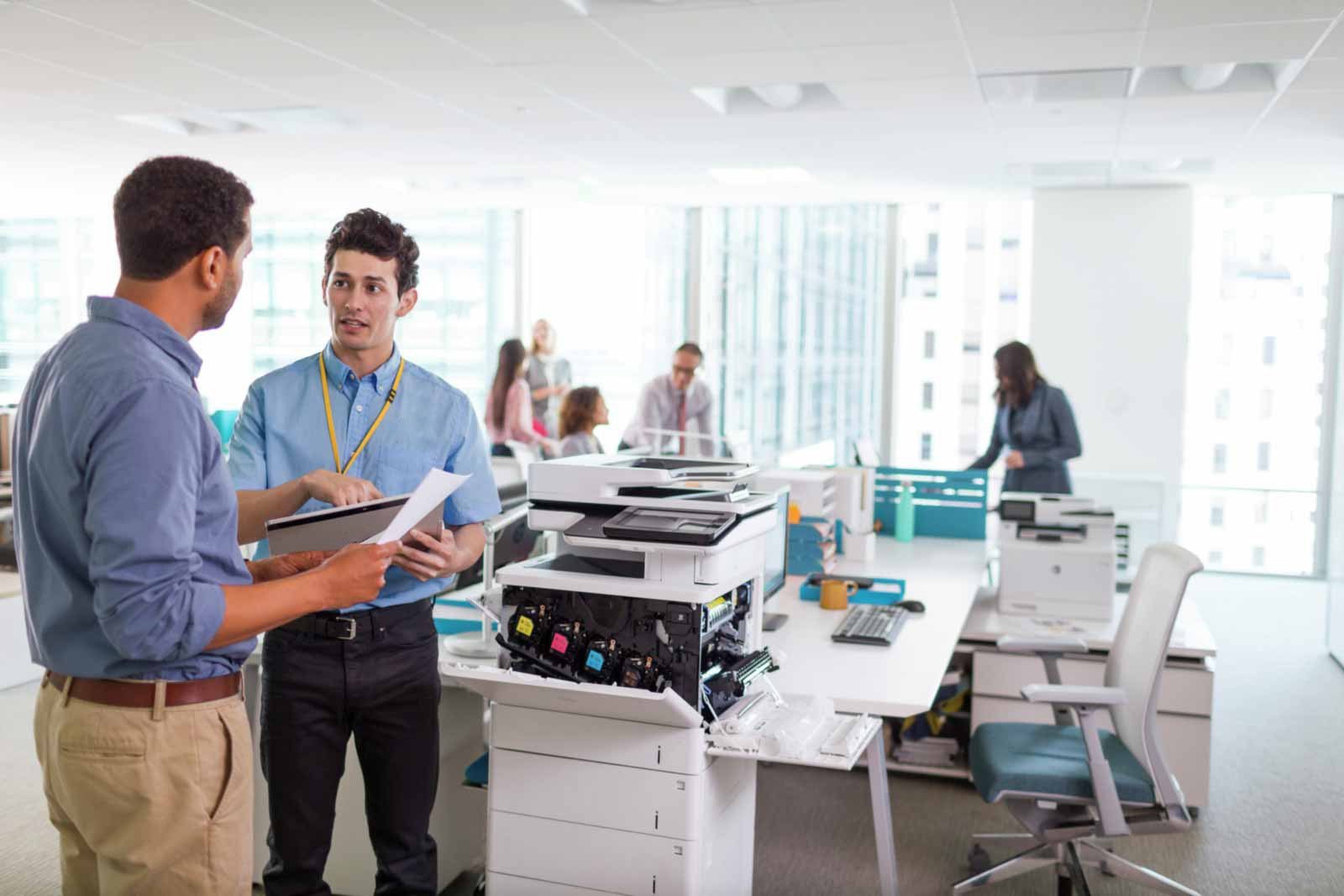 Two men in a modern office discuss a technical issue next to a large, open-paneled photocopier with visible colored ink cartridges.
